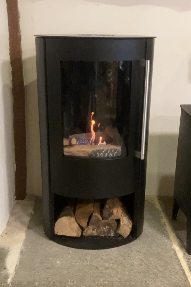 Black bio ethanol stove with a visible flame and log storage beneath, set on a stone hearth.
